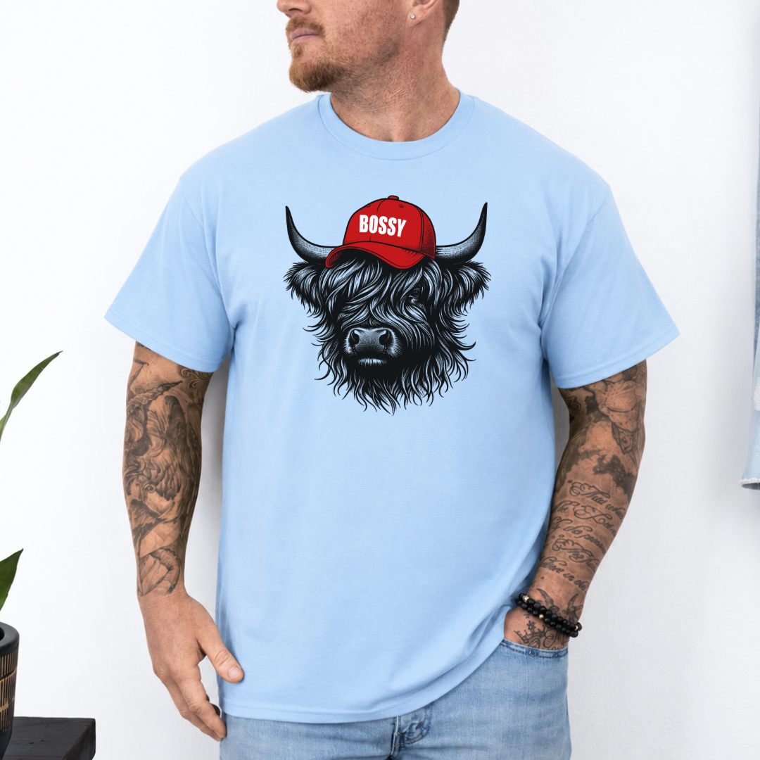 A man wearing a light blue t-shirt with a graphic of a black and white highland cow wearing a red cap that says 'BOSSY' in white letters. The man has tattoos on both arms and is wearing light wash jeans. Grab your bossy tee from Coffee and Moo NZ.