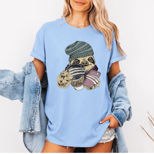 A woman wearing a light blue oversized graphic t-shirt with a cartoon sloth holding a coffee cup and a phone. She has a light-wash denim jacket off her shoulders and is posing against a clean white background. Grab your tee from Coffee and Moo NZ.