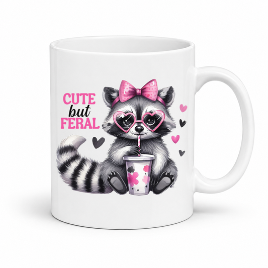 A white ceramic mug featuring a cute cartoon raccoon wearing pink heart-shaped glasses and a pink bow, holding a pink drink. The text 'CUTE but FERAL' is printed in pink and black next to the raccoon, with small pink and black hearts scattered around. from Coffee and Moo.