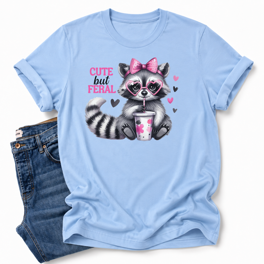 A light blue short-sleeve graphic T-shirt laid flat on a white background, styled with folded blue jeans. The shirt features a cartoon raccoon wearing a pink bow and heart-shaped glasses, holding a drink, with the text 'CUTE but FERAL' in pink and black. Grab your tee from Coffee and Moo NZ.