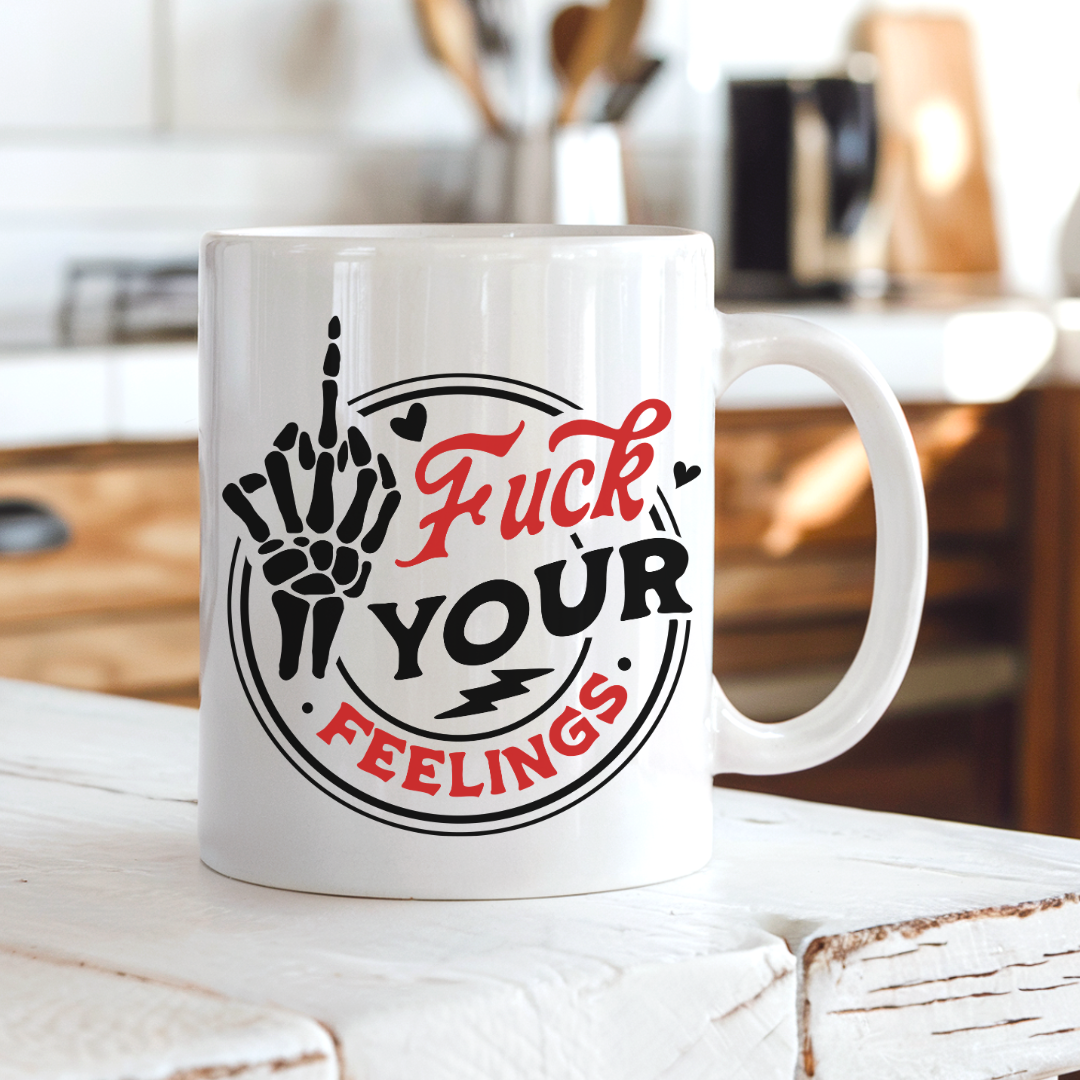 A white ceramic coffee mug with a bold graphic design, sitting on a rustic white wooden table in a warm, blurred kitchen setting. The graphic features a skeleton hand giving a middle finger in black, with the words 'Fuck YOUR FEELINGS' in red and black script. Purchase from Coffee and Moo NZ.