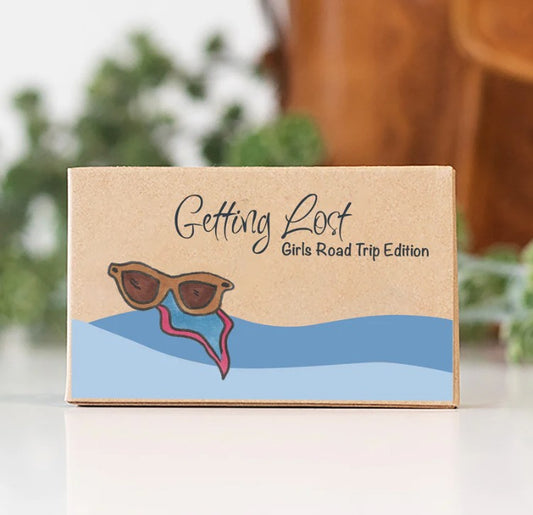 'Getting Lost Girls Road Trip Edition' game sits on a white surface with a blurred indoor background. The box is kraft brown with a printed illustration of sunglasses over blue waves and a red trail. Purchase yours from Coffee and Moo NZ