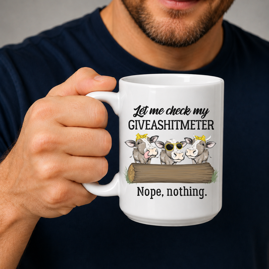 A person holding a white ceramic mug with a humorous graphic that reads 'Let me check my GIVEASHITMETER Nope, nothing.' above three cartoon cows behind a log. Grab your cheeky mug from Coffee and Moo NZ.