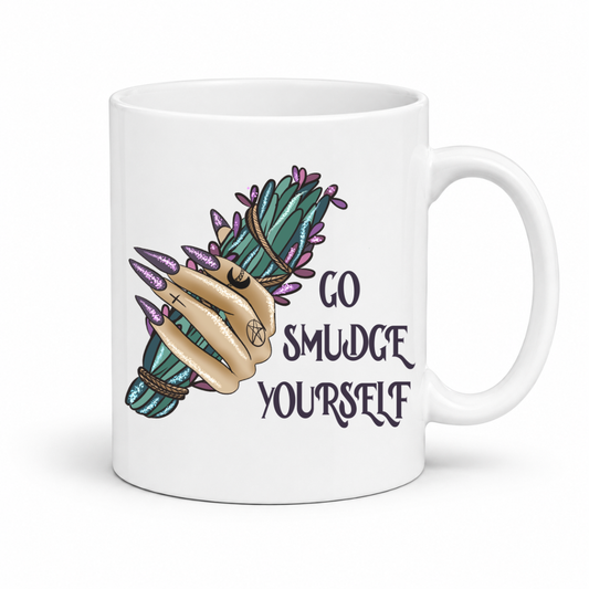 A white ceramic mug featuring a hand holding a bundle of purple and green herbs, with the text 'GO SMUDGE YOURSELF' printed in a dark gothic font. Purchase from Coffee and Moo NZ.
