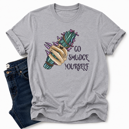 A grey short-sleeve t-shirt with a graphic of a hand holding a smudge stick and the text 'GO SMUDGE YOURSELF' in dark lettering. The t-shirt is styled flat-lay with dark blue jeans on a clean white background. Grab your tee from Coffee and Moo NZ.