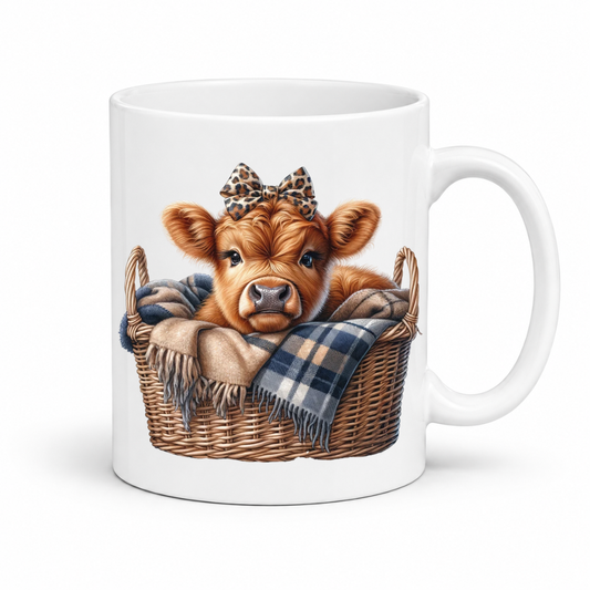 A white ceramic mug featuring a printed illustration of a brown cow calf sitting in a wicker basket with a plaid blanket and a leopard-print bow. from Coffee and Moo.