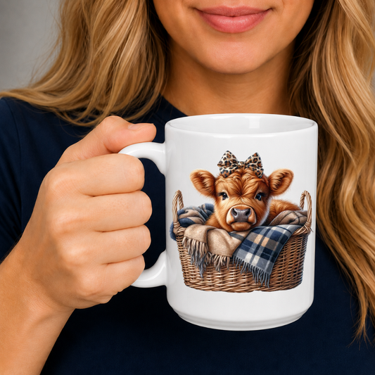 A person holding a white ceramic mug with a printed illustration of a brown cow calf in a basket with a plaid blanket and a leopard-print bow. The person is wearing a dark navy shirt. from Coffee and Moo.