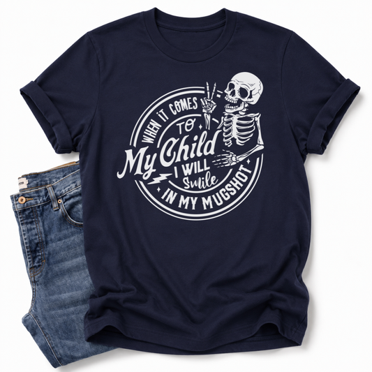 A navy blue short-sleeve t-shirt with a white graphic print of a skeleton and the text 'When it comes to My Child I Will Smile in my mugshot', styled flat-lay next to a pair of blue jeans on a white background. Grab your tee from Coffee and Moo NZ.