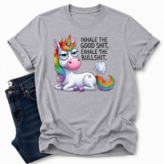 A grey short-sleeve t-shirt with a graphic of a cartoon unicorn and the text 'INHALE THE GOOD SHIT, EXHALE THE BULLSHIT.' styled flat-lay next to a pair of dark blue jeans on a white background. Grab your tee from Coffee and Moo NZ.