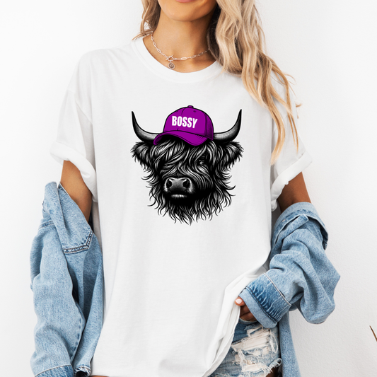 A woman wearing a white oversized graphic t-shirt with a black and white highland cow illustration wearing a purple cap that says 'BOSSY'. She has a light blue denim jacket draped off her shoulders and is posing against a plain white wall. Grab your Bossy highland coo tee from Coffee and Moo NZ.