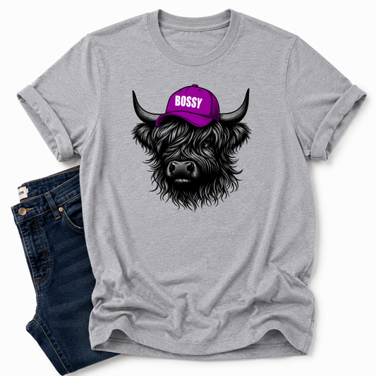 A heather gray short-sleeve t-shirt with a graphic of a black highland cow wearing a purple cap that says 'BOSSY' in white letters. The t-shirt is styled flat-lay with dark blue jeans on a white background. Grab your Bossy highland coo tee from Coffee and Moo NZ.