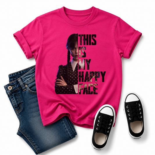 A bright pink graphic T-shirt laid flat on a white background, styled with folded blue jeans and a pair of black low-top sneakers. The shirt features a printed portrait of Wednesday Addams and the text 'THIS IS MY HAPPY FACE' in black. Grab your Kids tee from Coffee and Moo NZ