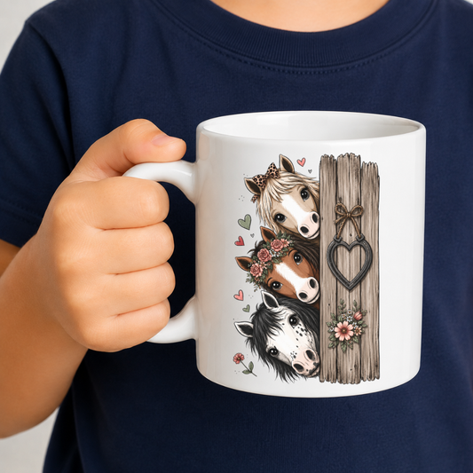 A child holding a white ceramic mug with a printed illustration of three horses peeking from behind a wooden fence, wearing a navy blue shirt. From Coffee and Moo