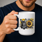 A person holding a white ceramic mug with a graphic print of a black French bulldog wearing a bandana next to a yellow sunflower, set against a dark navy blue shirt. Grab your frenchie mug from Coffee and Moo NZ