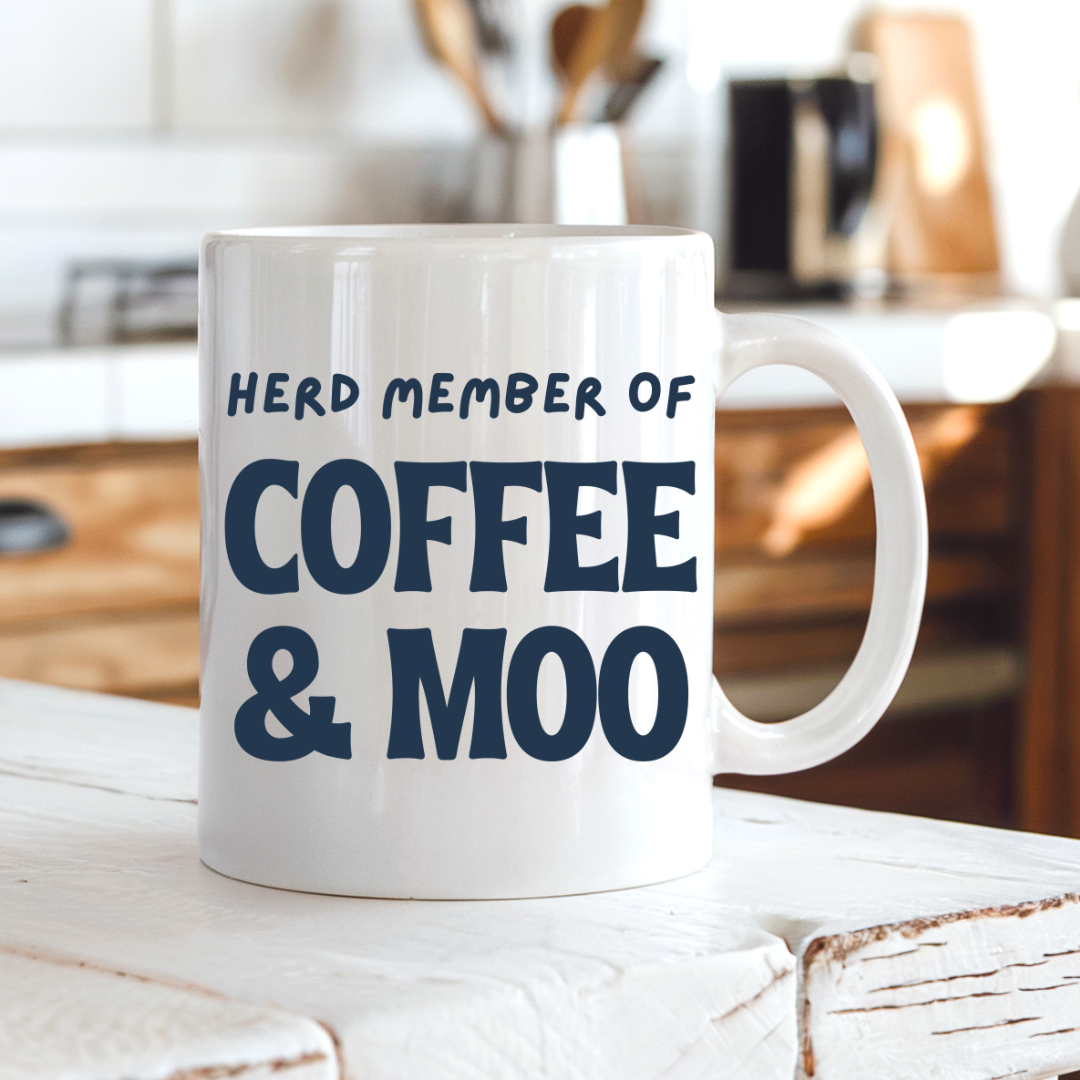 A white ceramic coffee mug with the text 'HERD MEMBER OF COFFEE & MOO' printed in dark navy blue, sitting on a rustic white wooden table in a warm, blurred kitchen setting. Grab your mug from Coffee and Moo NZ.