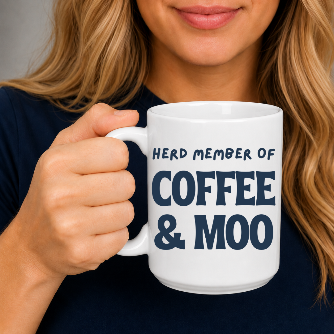 A woman holding a white ceramic coffee mug with the text 'HERD MEMBER OF COFFEE & MOO' printed in dark navy blue. The woman has blonde hair and is wearing a dark navy top, shown from the chest up against a neutral gray background. Grab your mug from Coffee and Moo NZ.
