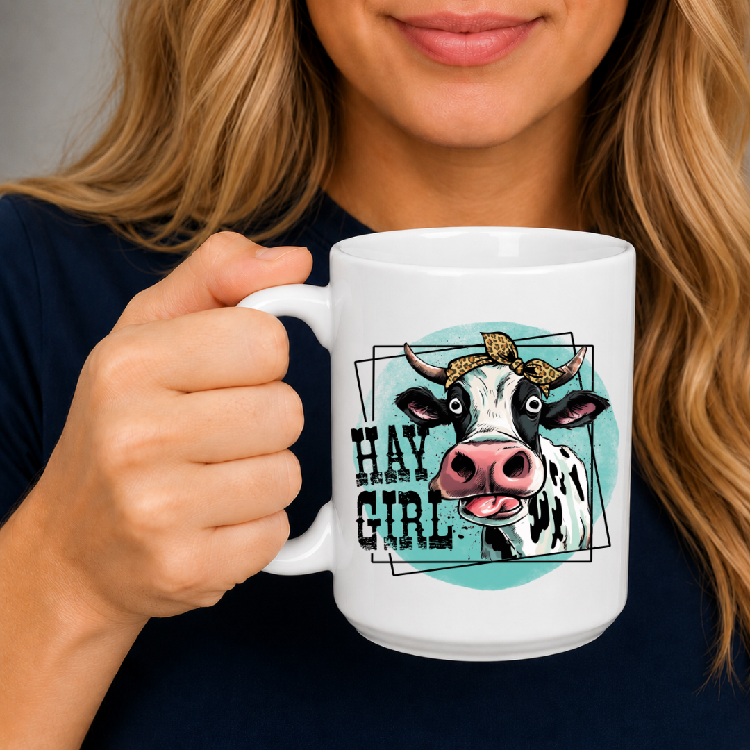 A woman holding a white ceramic mug with a graphic of a cow and the text 'HAY GIRL' on a dark navy shirt.  from Coffee and Moo NZ.