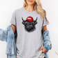 A woman wearing a grey t-shirt with a graphic of a black highland cow wearing a red cap that says 'BOSSY' in white. She has a light blue denim jacket draped off her shoulders against a plain white background. Grab your bossy tee from Coffee and Moo NZ.