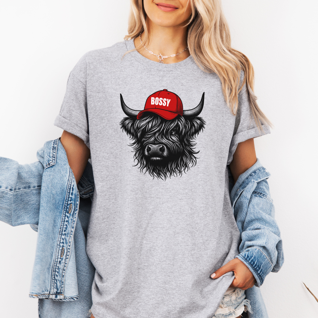A woman wearing a grey t-shirt with a graphic of a black highland cow wearing a red cap that says 'BOSSY' in white. She has a light blue denim jacket draped off her shoulders against a plain white background. Grab your bossy tee from Coffee and Moo NZ.