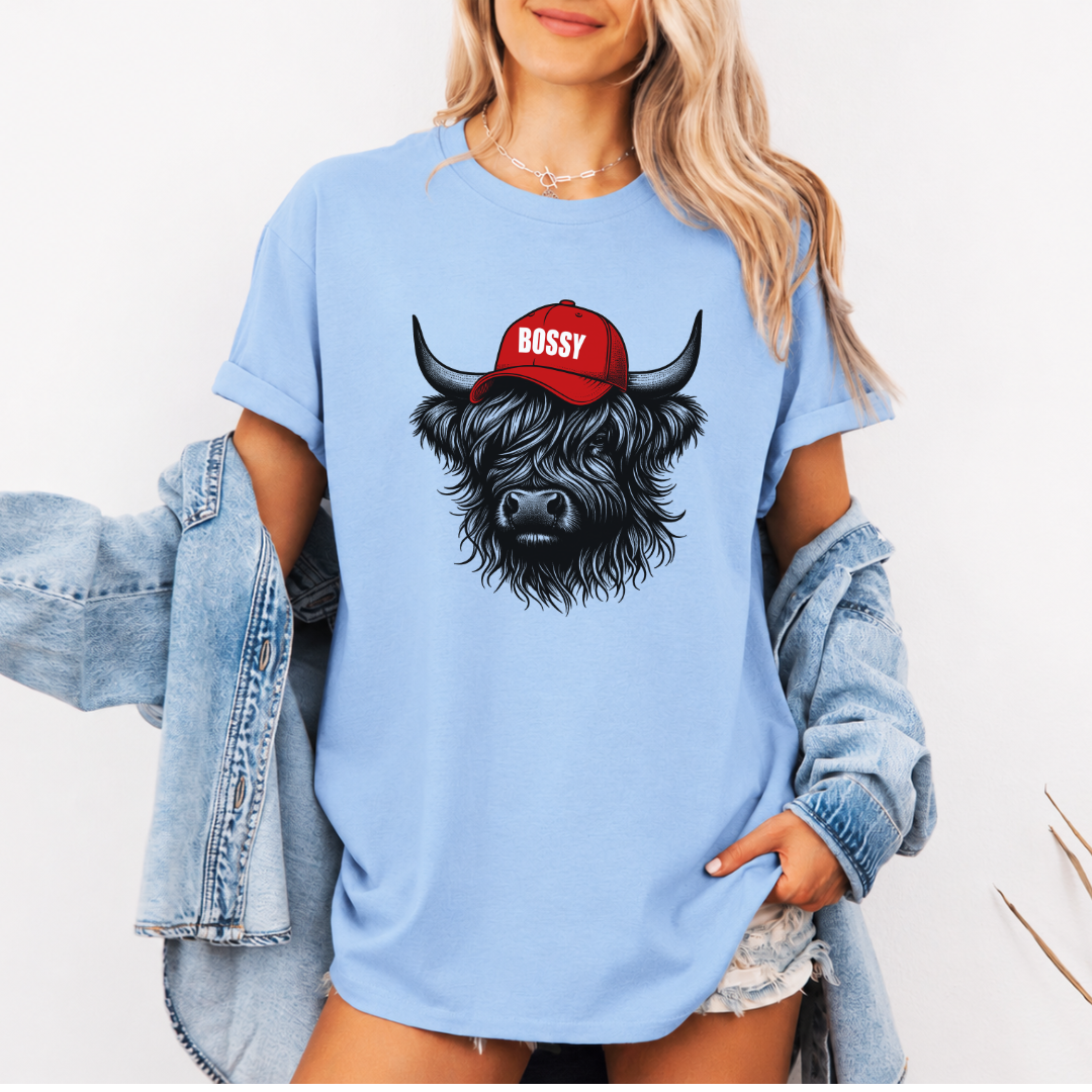 A woman wearing a light blue oversized graphic t-shirt with a black highland cow illustration wearing a red cap that says 'BOSSY' in white. She has a light wash denim jacket off her shoulders and is posing against a clean white background. Grab your bossy tee from Coffee and Moo NZ.
