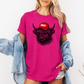 A woman wearing a bright pink graphic t-shirt with a black highland cow illustration wearing a red cap that says 'BOSSY' in white. She has a light blue denim jacket draped off her shoulders and is standing against a plain white wall. Grab your bossy tee from Coffee and Moo NZ.