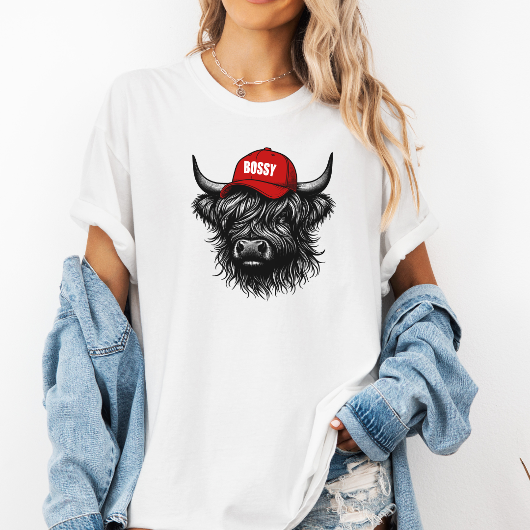 A woman wearing a white oversized graphic t-shirt with a black and red illustration of a Scottish Highland cow wearing a red cap that says 'BOSSY'. She has a light blue denim jacket off her shoulders and distressed jeans against a plain white background. Grab your bossy tee from Coffee and Moo NZ.