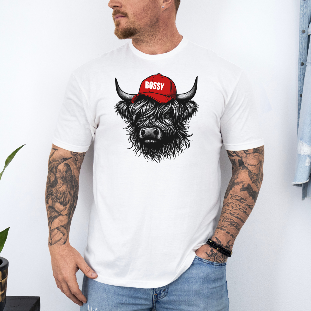 A man wearing a white t-shirt with a graphic of a black Scottish highland cow wearing a red cap that says 'BOSSY' in white text, paired with blue jeans. Grab your bossy tee from Coffee and Moo NZ.