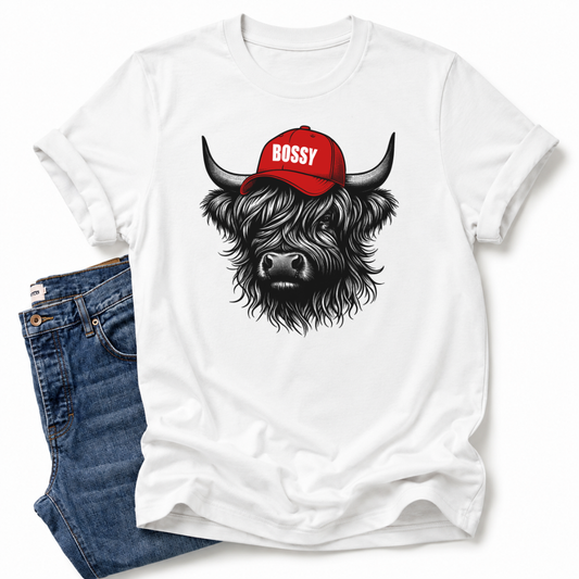 A white short-sleeve T-shirt laid flat with a graphic of a shaggy black-and-gray Highland cow wearing a red cap with the word 'BOSSY' in white. Blue jeans are partially visible underneath on the left side. Grab your bossy tee from Coffee and Moo NZ.