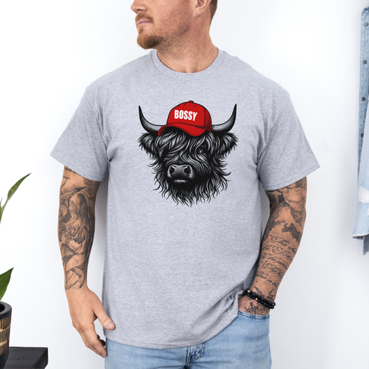 A man wearing a grey t-shirt with a graphic of a black highland cow wearing a red cap that says 'BOSSY' in white text. The man is also wearing light blue jeans, standing against a white wall. Grab your bossy tee from Coffee and Moo NZ.