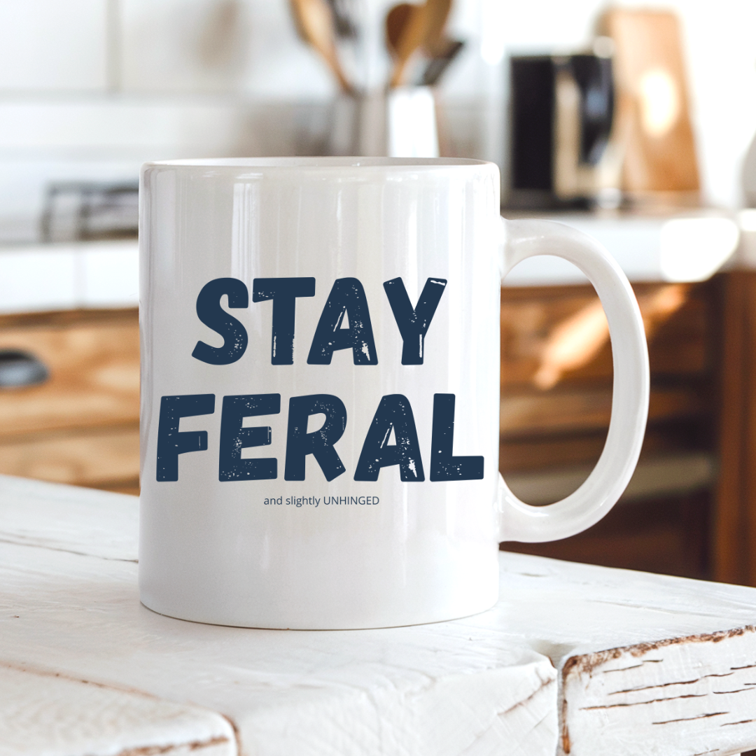 A white ceramic coffee mug with the text 'STAY FERAL and slightly UNHINGED' printed in navy blue, sitting on a rustic white wooden table in a warm, blurred kitchen setting. Grab your feral mug from Coffee and Moo NZ.