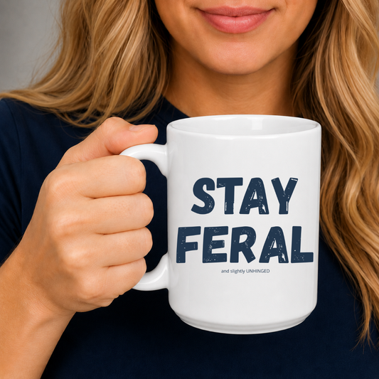 A woman holding a white ceramic mug with the text 'STAY FERAL and stylish UNHINGED' printed in dark navy blue. The woman has blonde hair and is wearing a dark navy top, shown from the chest up against a neutral grey background. Grab your feral mug from Coffee and Moo NZ.