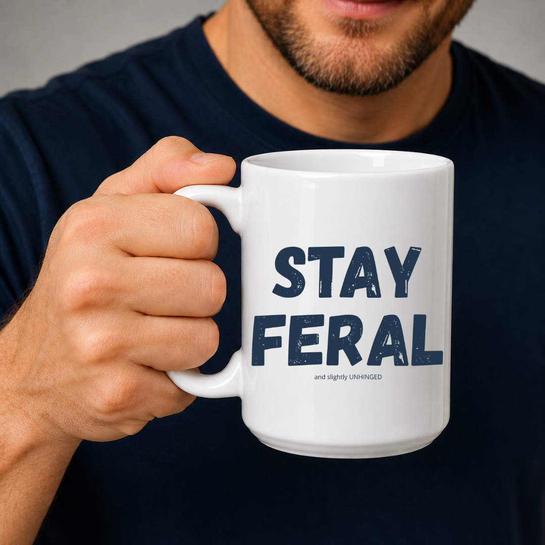 A person in a navy blue shirt holds a white ceramic mug with the text 'STAY FERAL' and smaller text 'and aging over time' printed in dark navy lettering. Grab your feral mug from Coffee and Moo NZ.