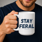 A person in a navy blue shirt holds a white ceramic mug with the text 'STAY FERAL' and smaller text 'and aging over time' printed in dark navy lettering. Grab your feral mug from Coffee and Moo NZ.