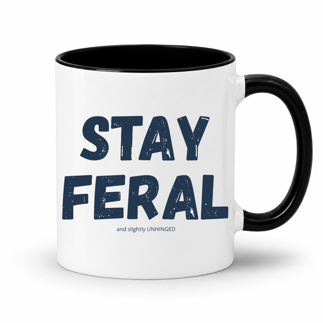 A white ceramic mug with a black interior, black handle, and navy blue text reading 'STAY FERAL' with smaller text 'and slightly UNHINGED' underneath, shown on a clean white background. Grab your feral mug from Coffee and Moo NZ.