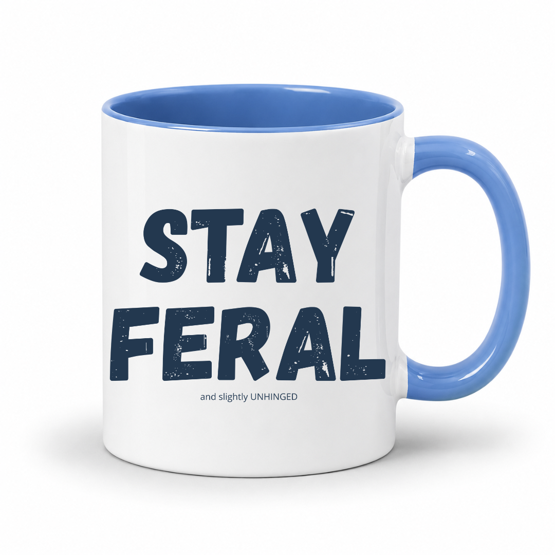 A white ceramic coffee mug with a blue interior, blue handle, and navy blue text reading 'STAY FERAL' with smaller text 'and slightly UNHINGED' underneath, shown on a white background. Grab your feral mug from Coffee and Moo NZ.