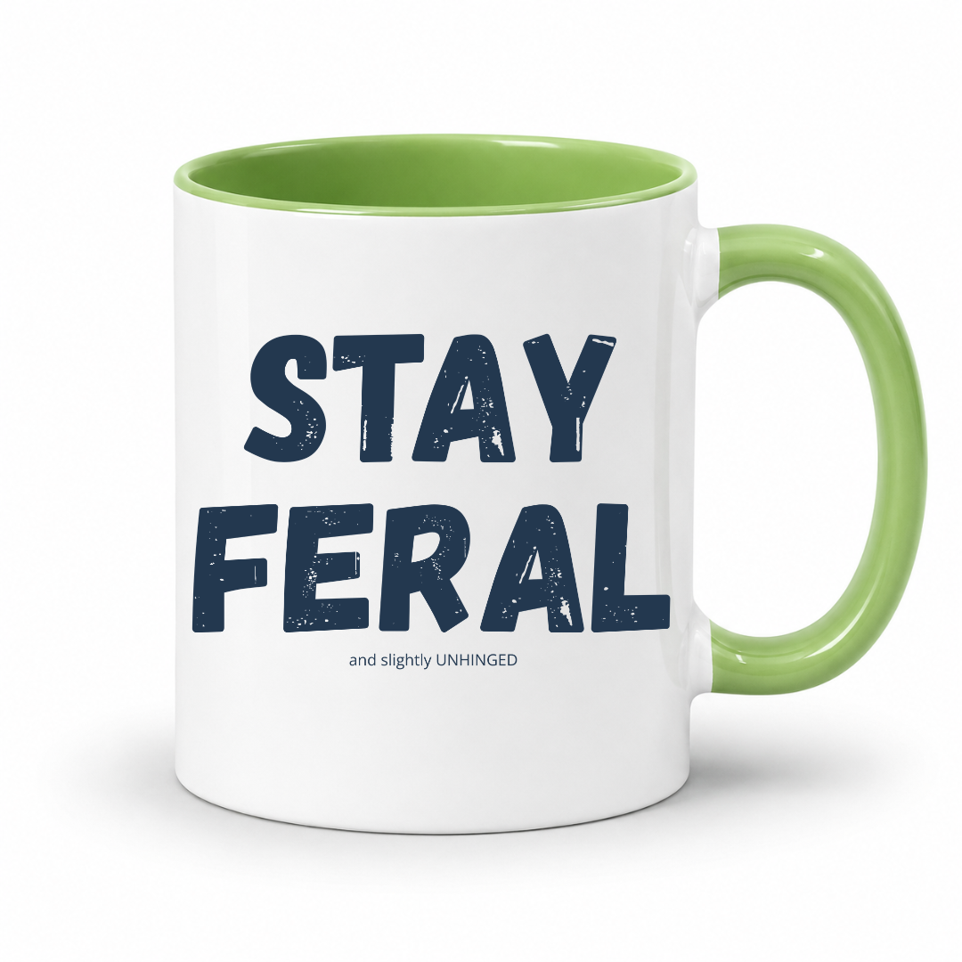A white ceramic coffee mug with a light green interior and handle, featuring the text 'STAY FERAL' in dark navy blue with a small tagline underneath, on a clean white background. Grab your feral mug from Coffee and Moo NZ.