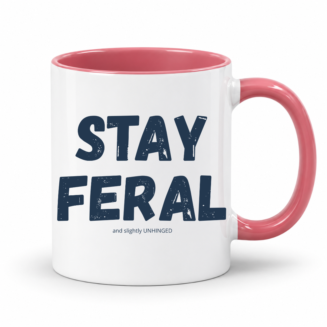 A white ceramic coffee mug with a pink interior and handle, featuring the text 'STAY FERAL' in dark navy blue letters and 'and slightly UNHINGED' in smaller text below, on a white background. Grab your feral mug from Coffee and Moo NZ.