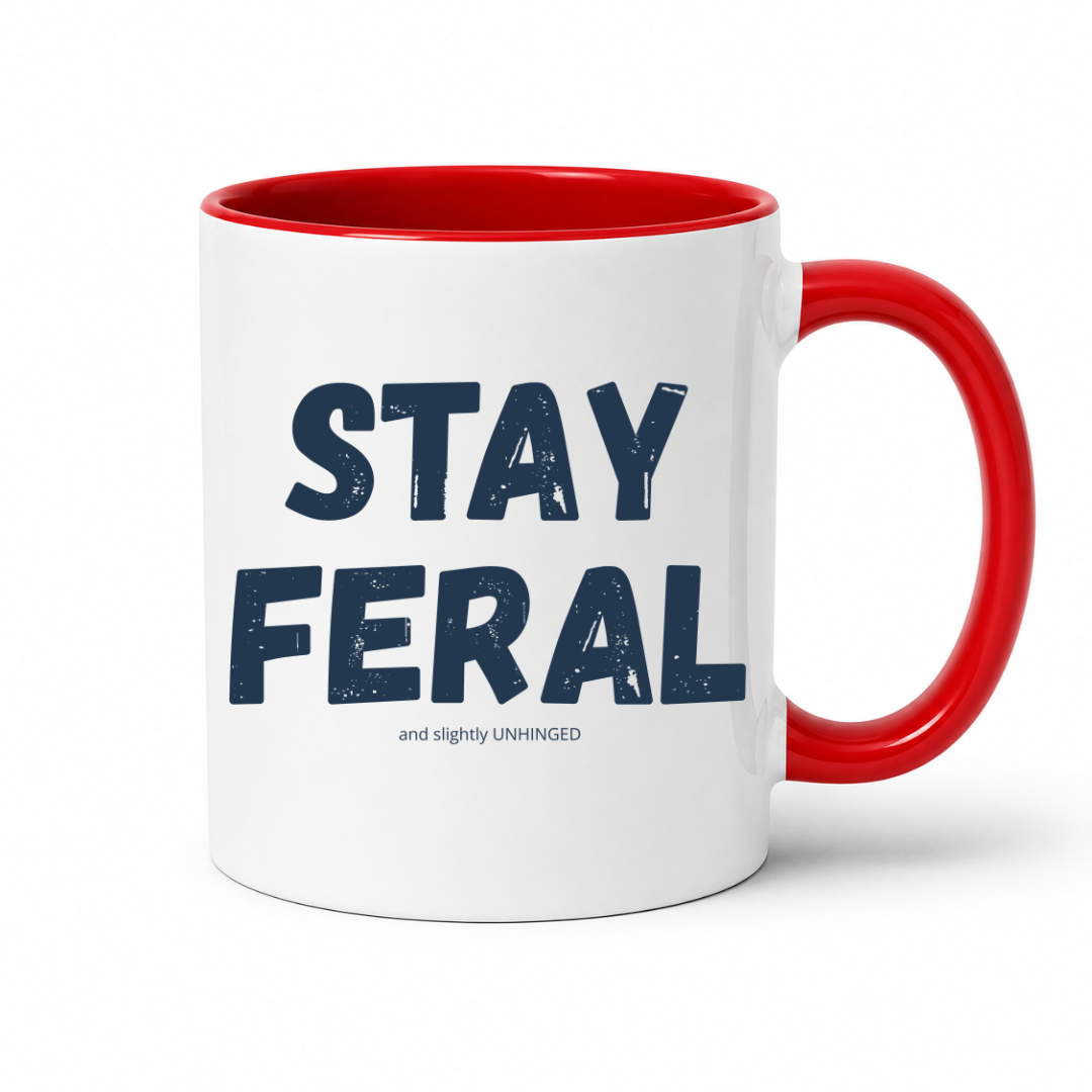 A white ceramic coffee mug with a red interior and red handle, featuring the text 'STAY FERAL' in dark navy blue letters and a smaller tagline underneath, set against a plain white background. Grab your feral mug from Coffee and Moo NZ.