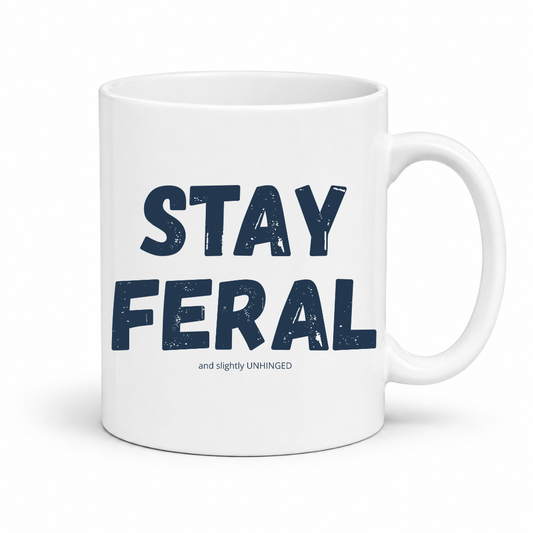 A white ceramic coffee mug with the words 'STAY FERAL' printed in a dark navy blue distressed font, with a smaller tagline 'and slightly UNHINGED' underneath, shown on a clean white background. Grab your feral mug from Coffee and Moo NZ.