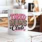A white ceramic mug with the phrase 'TIRED MUMS Club' printed on it, featuring pink lettering and leopard print details, set on a rustic white wooden table with a blurred kitchen background. Grab your mug from Coffee and Moo NZ.
