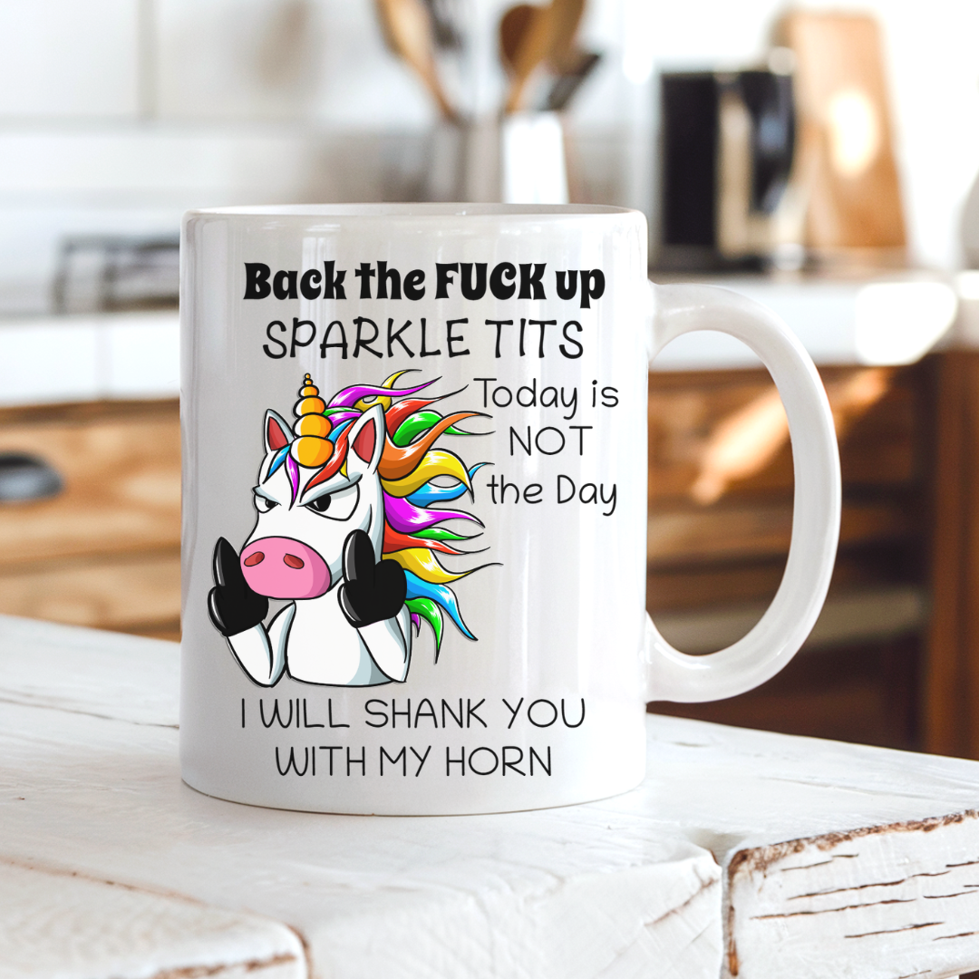 A white ceramic mug with a humorous graphic of a cartoon unicorn and the text 'Back the FUCK up SPARKLE TITS Today is NOT the Day I WILL SHANK YOU WITH MY HORN', shown on a rustic white wooden table with a blurred kitchen background. Grab yours from Coffee and Moo.