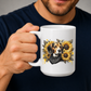 A person holding a white ceramic mug with a detailed graphic of a beagle dog surrounded by yellow sunflowers and a black bandana. The person is wearing a dark navy blue t-shirt against a neutral grey background. Grab your mug from Coffee and Moo NZ.