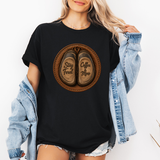 A woman wearing a black graphic t-shirt with a circular design of two cow hooves reading 'Stay Feral' and 'Coffee & Moo'. She has a light blue denim jacket off her shoulders and is wearing denim shorts against a plain white background. Grab your official Coffee & Moo brand tee from Coffee and Moo NZ.