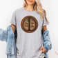 A woman wearing a grey graphic t-shirt with a circular design of two cow hooves reading 'Stay Feral' and 'Coffee & Moo'. She has a light blue denim jacket off her shoulders and is wearing denim shorts against a plain white background. Grab your official Coffee & Moo brand tee from Coffee and Moo NZ.