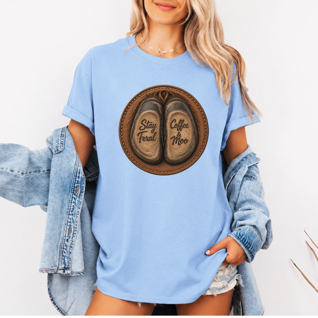 A woman wearing a baby blue graphic t-shirt with a circular design of two cow hooves reading 'Stay Feral' and 'Coffee & Moo'. She has a light blue denim jacket off her shoulders and is wearing denim shorts against a plain white background. Grab your official Coffee & Moo brand tee from Coffee and Moo NZ.