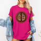 A woman wearing a pink graphic t-shirt with a circular design of two cow hooves reading 'Stay Feral' and 'Coffee & Moo'. She has a light blue denim jacket off her shoulders and is wearing denim shorts against a plain white background. Grab your official Coffee & Moo brand tee from Coffee and Moo NZ.