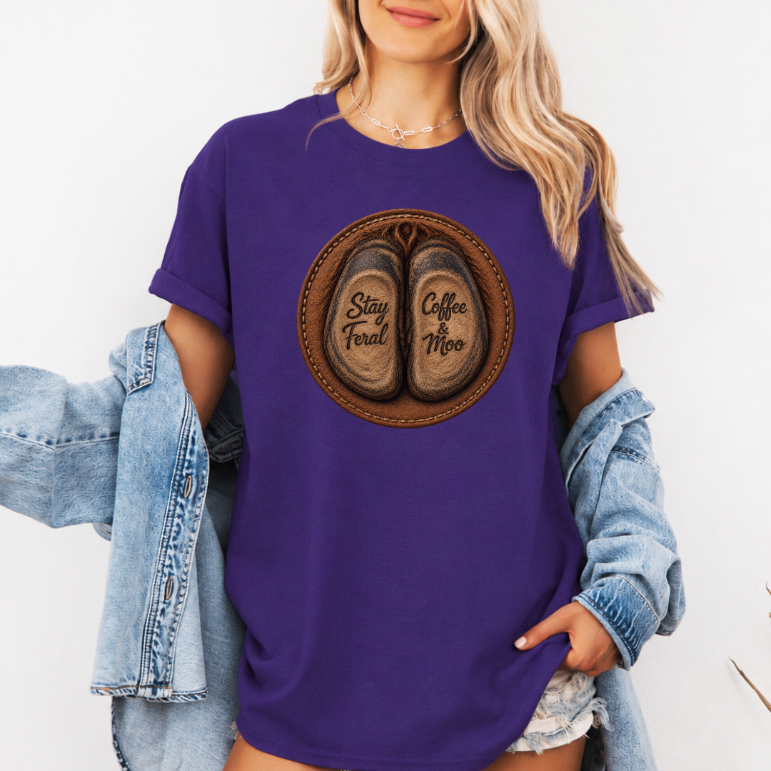 A woman wearing a purple graphic t-shirt with a circular design of two cow hooves reading 'Stay Feral' and 'Coffee & Moo'. She has a light blue denim jacket off her shoulders and is wearing denim shorts against a plain white background. Grab your official Coffee & Moo brand tee from Coffee and Moo NZ.