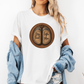 A woman wearing a white graphic t-shirt with a circular design of two cow hooves reading 'Stay Feral' and 'Coffee & Moo'. She has a light blue denim jacket off her shoulders and is wearing denim shorts against a plain white background. Grab your official Coffee & Moo brand tee from Coffee and Moo NZ.