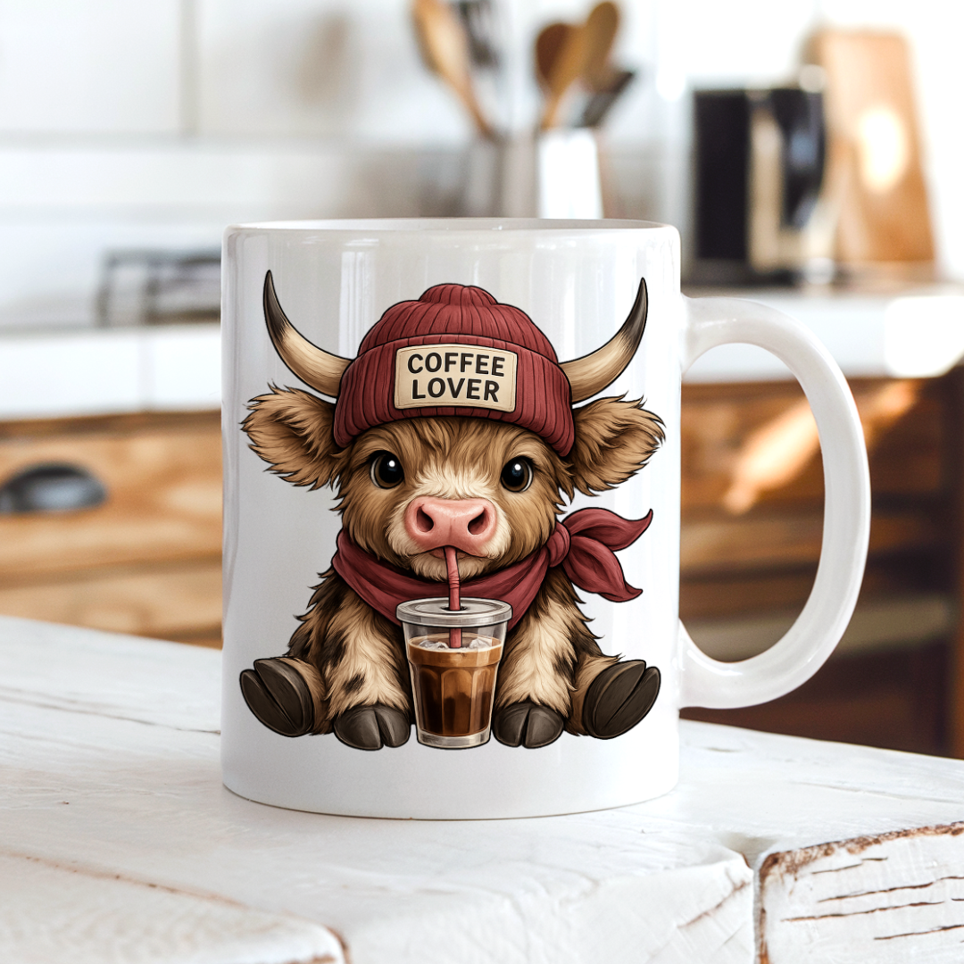 Mug with a cartoon highland cow wearing a 'Coffee Lover' hat and drinking from a cup on a kitchen counter. from Coffee and Moo.