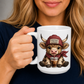 A person holding a white ceramic mug with a cartoon illustration of a cow wearing a maroon 'COFFEE LOVER' hat and scarf, sipping from a cup. The person is wearing a dark navy shirt against a neutral gray background from Coffee and Moo.